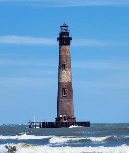 Morris Island Lighthouse standing tall above the surf under a blue sky.