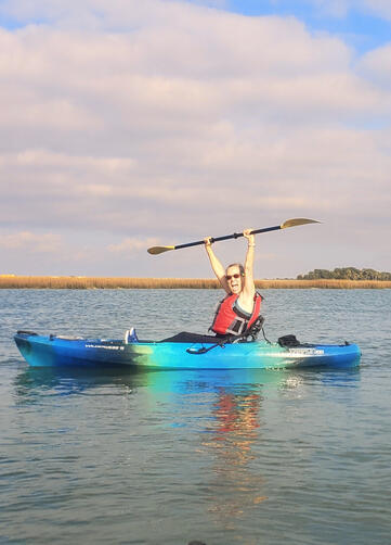 Person excitedly kayaking on calm water under a pastel sky.